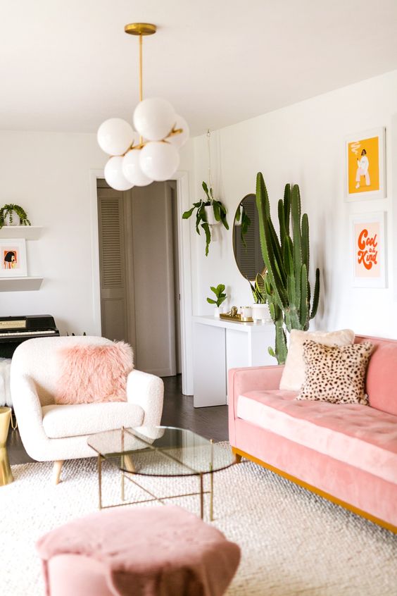 a pretty modern living room with a pink sofa, a stool and a faux fur pillow, potted plants, a retro chandelier and a glass table