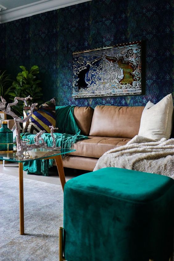 a sophisticated living room with a dark wallpaper wall, a brown sofa, emerald touches and a glass table plus potted plants