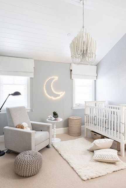 a beautiful neutral nursery with grey walls, grey and white furniture, printed pillows, a cool chandelier and a half moon lamp