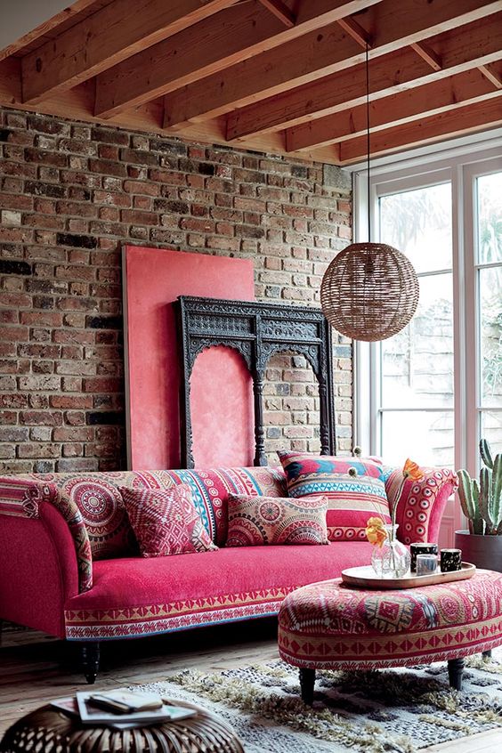 a boho living room with a brick wall, a pink sofa with printed pillows, a matching ottoman, a rattan pendant lamp and a Moroccan rug