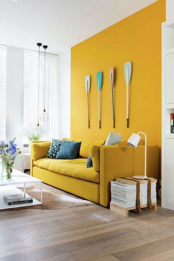a bold space with a mustard wall and a matching sofa, oars for decor, a low table and stacked magazines is quirky