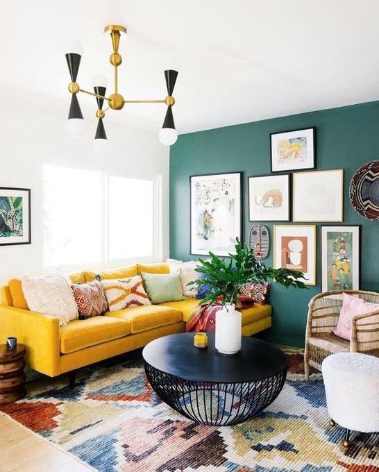 a bright boho living room with a green wall and a gallery wall, a yellow sectional, a retro chandelier and a black round table