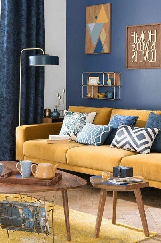 a chic living room with a navy accent wall, a yellow sofa with blue pillows, a yellow rug, a floor lamp and wooden tables