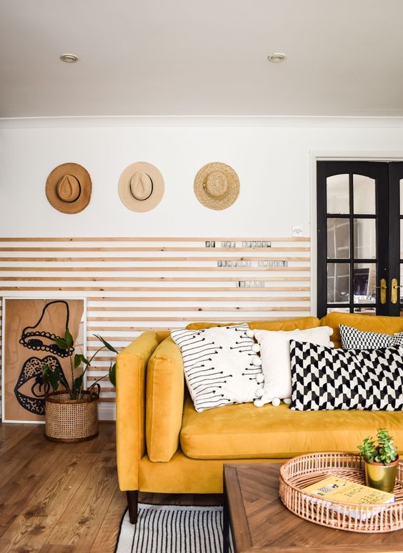 a chic living room with wooden slabs on the wall, a yellow sofa and graphic pillows, hats on display and some plants
