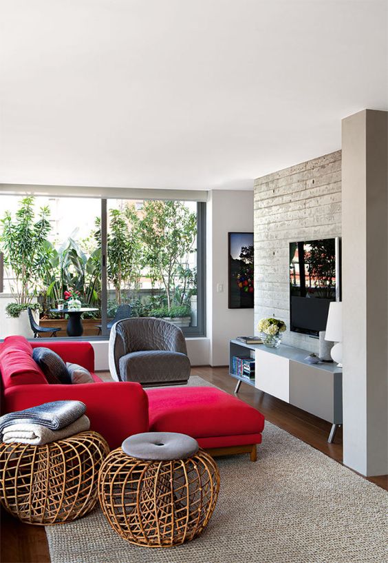a chic modern living room with a concrete wall, a two tone sideboard, a bold red sectional, a grey chair and rattan ottomans