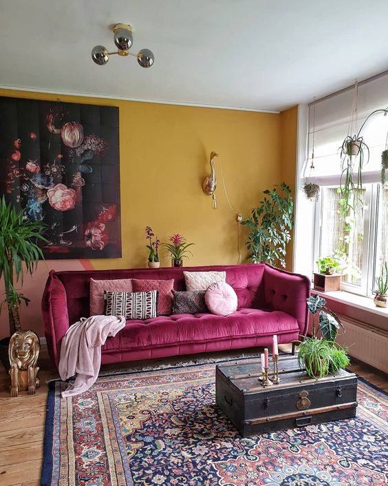 a chic modern living room with a mustard accent wall, a pink sofa, a boho printed rug, a vintage suitcase and potted plants