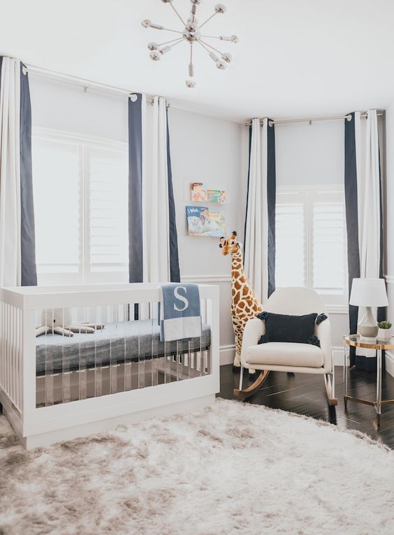 a chic modern nursery with a wood and acrylic crib, a rocker chair, an acrylic table, striped curtains and a dusty pink rug