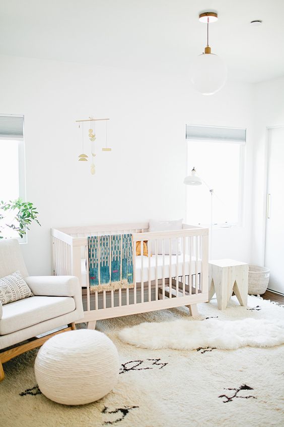 a chic neutral nursery with white furniture, layered rugs, a pouf, a mobile and some lamps is airy and serene