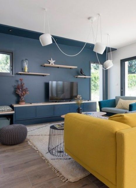 a contemporary living room with a navy accent wall, a yellow sofa, a grey chair, pendant lamps and floating shelves is chic