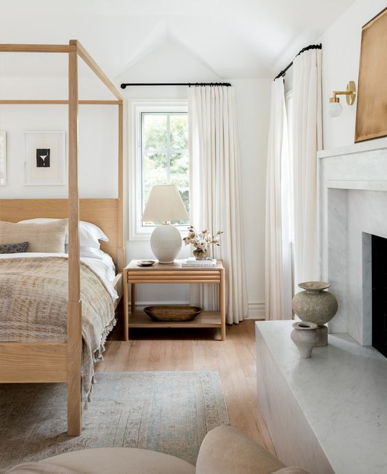 a cool and serene bedroom in neutrals, with a non-working fireplace, a blonde wood canopy bed and a matching nightstand is chic