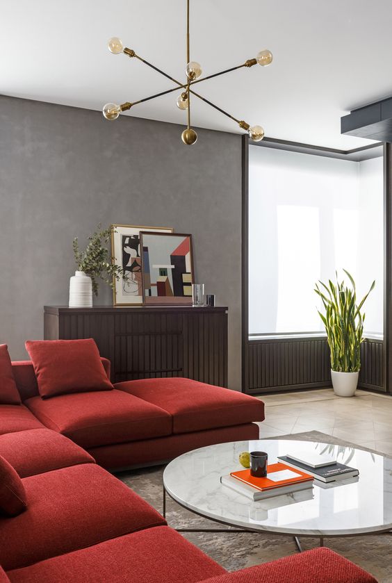 a laconic modern living room with grey walls, a depe red sectional, a wooden storage unit and a low round table