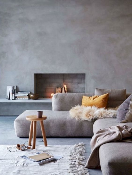 a lovely minimalist living room with concrete walls, a built-in fireplace, a low grey sofa, a roudn stool and various textiles