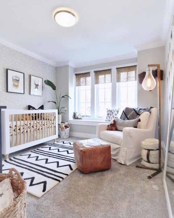 a lovely neutral nursery with a large crib, a white chair and a leather pouf, layered rugs, a basket and a floor lamp plus a grey polka dot wall