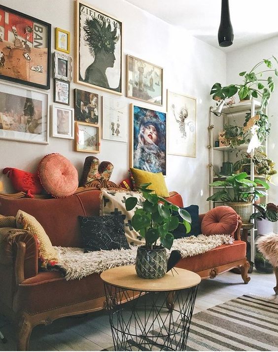 A maximalist living room with a colorful gallery wlal, a rust colored vintage sofa, a round table and potted plants
