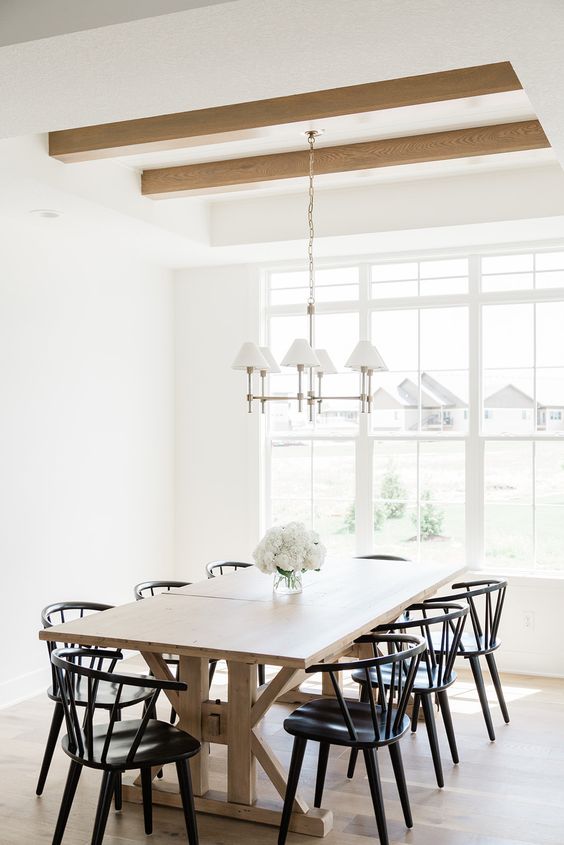 A mid century modern dining room with a blonde wood trestle table and beams, black chairs and a vintage chandelier