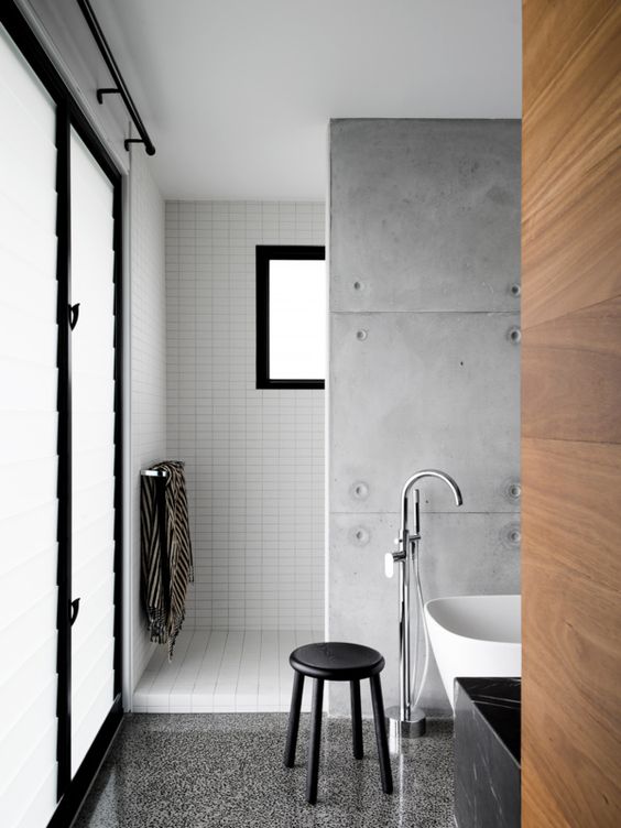 a minimalist industrial bathroom with white skinny tiles, a concrete tile wall and a grey terrazzo floor plus a white bathtub