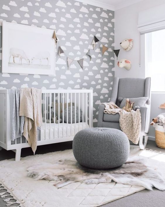 a modern grey nursery with a cloud print wlal, a grey rocker and a white crib, a grey pouf, a bunting and faux taxidermy for a fun touch