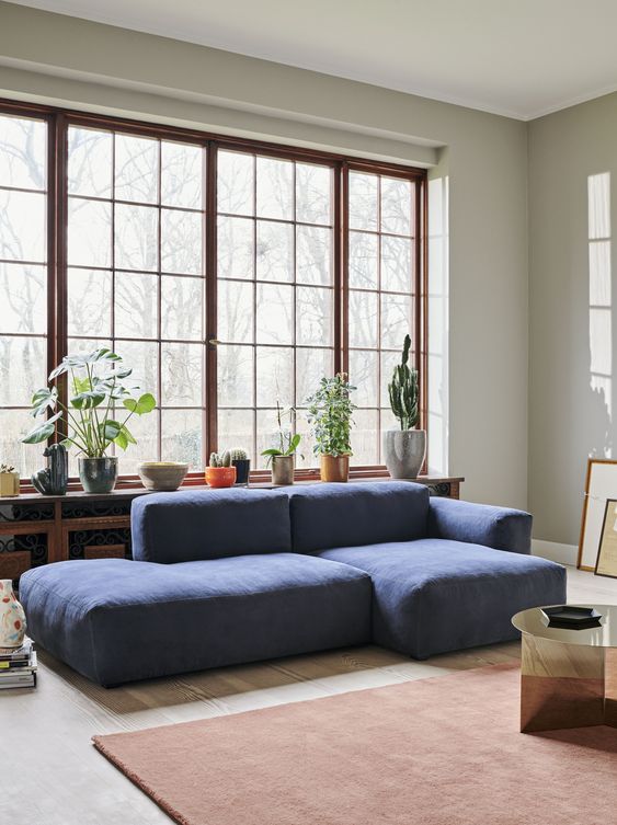 a modern living room with a low blue sofa, a metallic table, a rust rug and a whole collection of potted plants on the windowsill