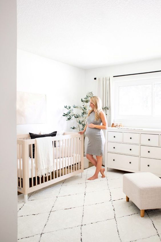 a modern neutral nursery with a light stained crib, a white dresser and a neutral ottoman plus neutral textiles