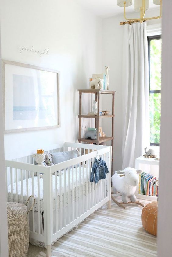 a modern neutral nursery with a white crib, some storage units, a leather pouf, some toys and a chic chandelier