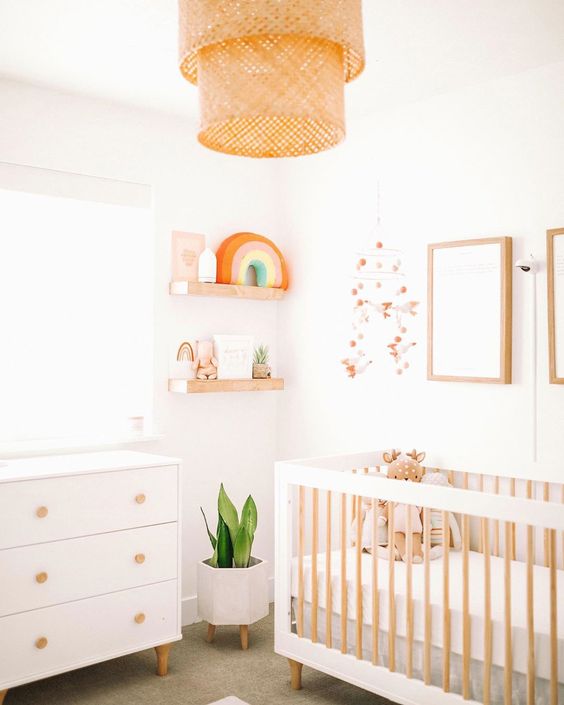 a modern neutral nursery with a white dresser and a large crib, a woven lamp and shelves with pretty decor