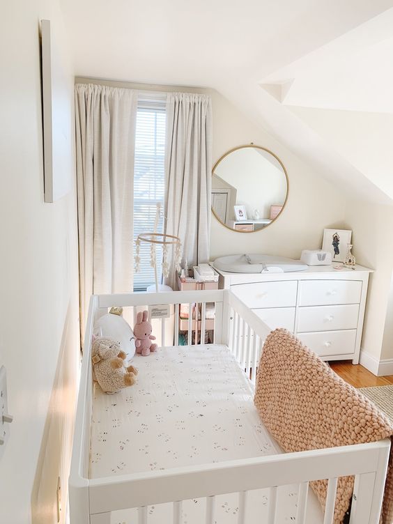 a modern neutral nursery with white furniture, printed and pastel bedding, grey curtains, a round mirror and pretty toys