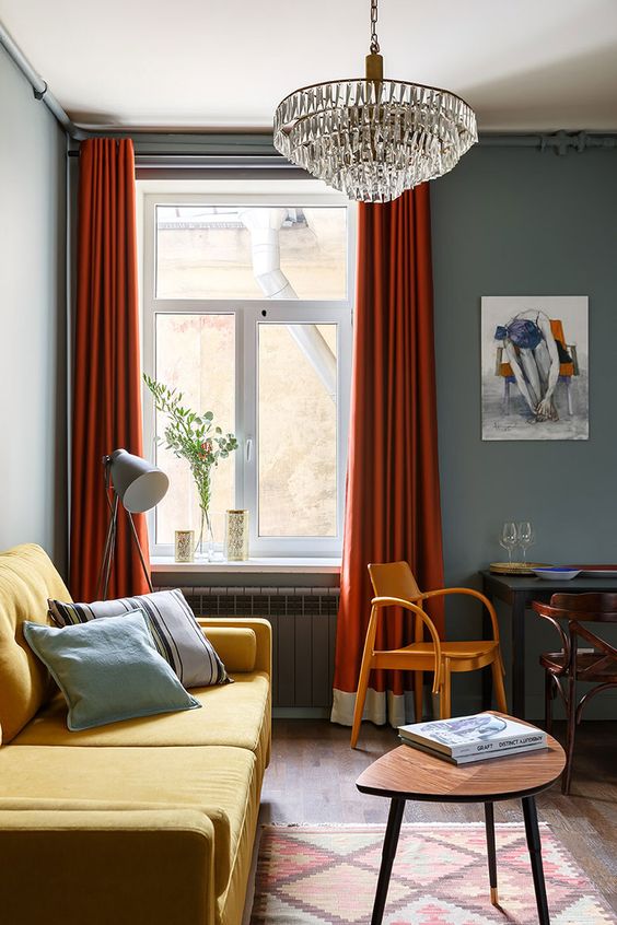 A moody living room with grey walls, a yellow sofa, rust colored curtains, a crystal chandelier and vintage chairs and tables