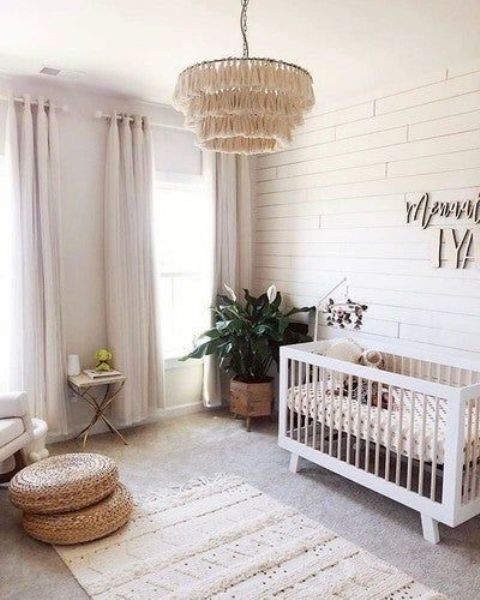 a neutral boho nursery with a white crib, a chair and a small table, jute cushions, a boho rug and a tassel chandelier