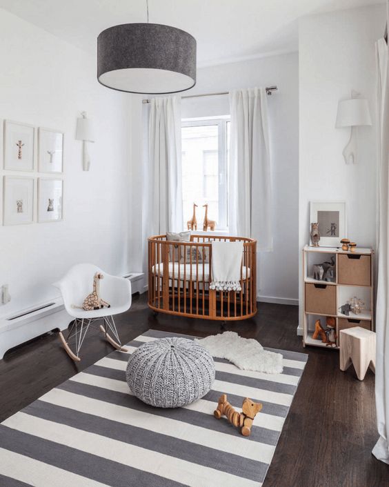 a neutral nursery with a stained crib, a white chair and a stool, a storage unit, a striped rug, a pendant lamp and a pouf