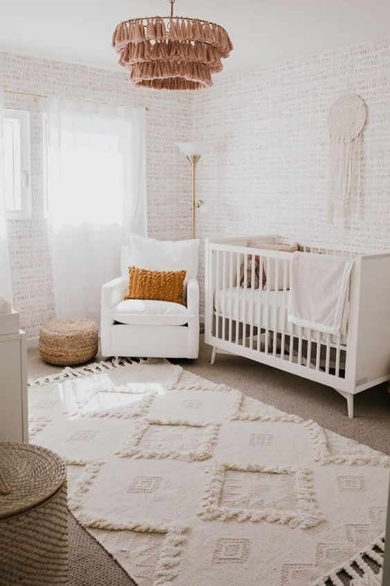 a neutral nursery with white furniture, a basket for storage, a tassel chandelier, a jute pouf, boho rugs