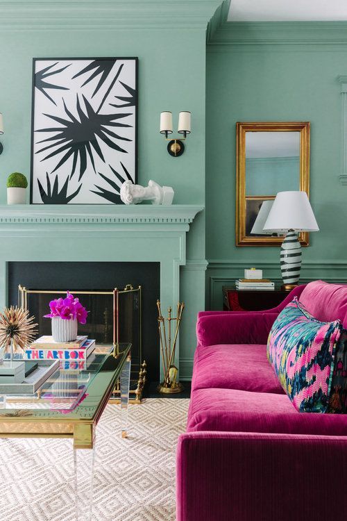 a pastel green living room with a non-working fireplace, lovely artworks, a fuchsia sofa with florla pillows and a lovely table