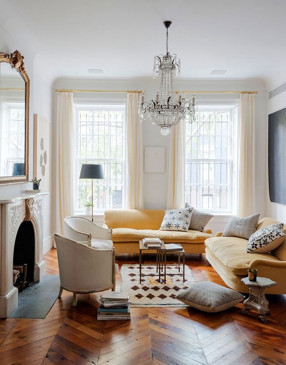 A refined Parisian living room with a non working fireplace, a yellow sectional, a refined crystal chandelier, creamy chairs and a large mirror over the fireplace