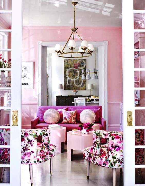 a refined feminine living room with pink walls, a pink loveseat, floral chairs, blush coffee tables and pillows and a chic chandelier