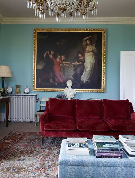 a refined living room with blue walls, a deep red sofa, a floral ottoman, a bold vintage artwork and a crystal chandelier
