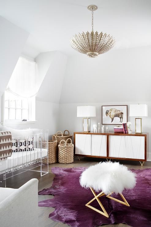 a refined modern nursery with neutral walls, a white sideboard, an acrylic crib, a faux fur stool and a pink rug plus a glam chandelier