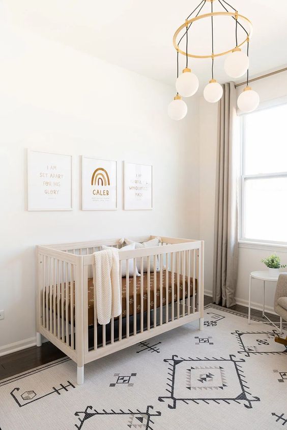 a serene and neutral nursery with a printed rug, grey curtains, a neutral crib and a pretty gallery wall plus a chandelier with hanging bulbs