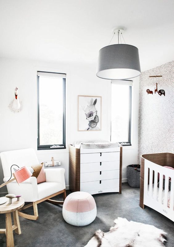 a stylish modern nursery with black framed windows, a grey chandelier, a white rocker and white and stained furniture, a colro block pouf and layered rugs