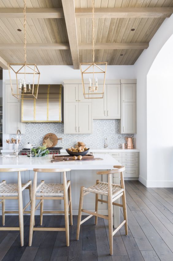 a very chic kitchen in light grey, with a blonde wood ceiling and tall stools with woven seats, gold pendant lamps