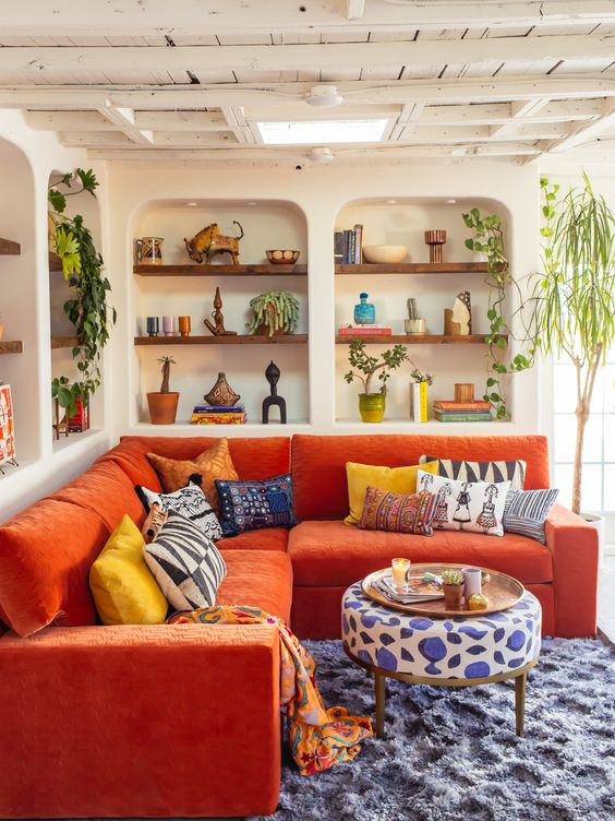 A vivacious living room with niches with built in shelves, an orange sofa, bold textiles and potted plants for a maximalist feel