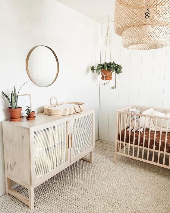 a welcoming neutral nursery with a jute rug, a crib, a rattan credenza, a woven lamp and a round mirror plus potted plants