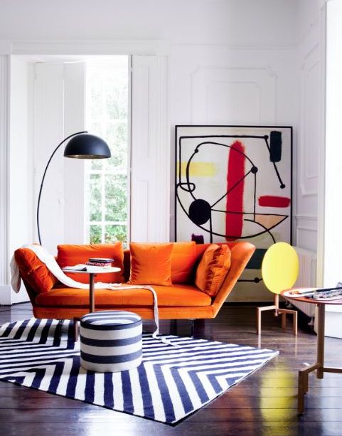 a whimsy living room with an orange loveseat, a bold abstract artwork, a floor lamp and a geometric rug and a pouf