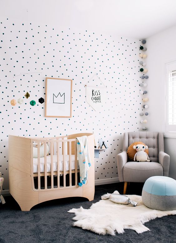 a whimsy modern nursery with a polka dot accent wall, a grey chair, a light stained crib, layered rugs and a color block pouf and cool lights
