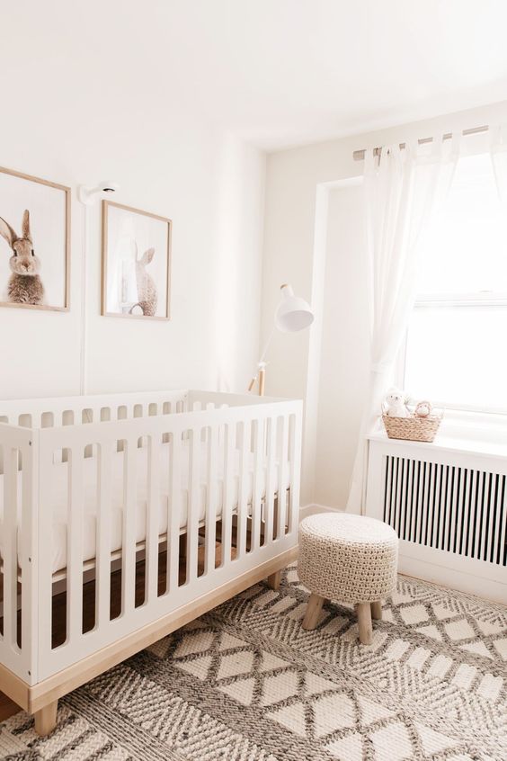 an airy neutral nursery with a white crib and a woven stool, a boho rug, a basket and a pretty and cute gallery wall