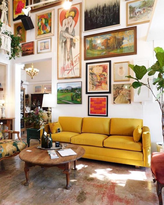 an artful living room with a bold gallery wall coming up to the ceiling, a yellow sofa, a vintage wooden table and a printed chair