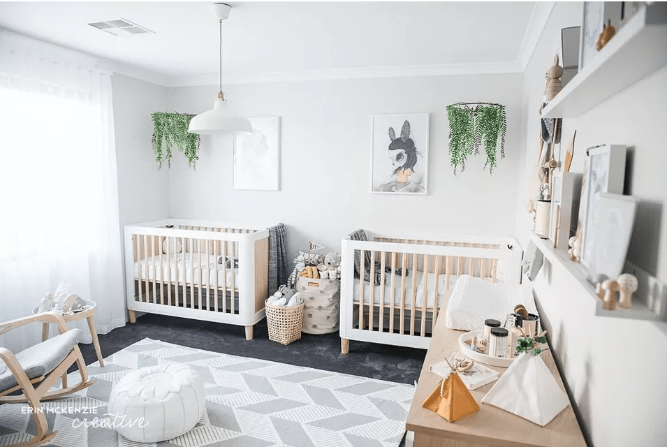 a beautiful and fresh twin nursery with white cribs and a chair, a round table and a stained dresser, greenery mobiles and pretty artworks
