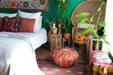 a boho maximalist bedroom with green walls, a colorful rug, a peacock chair, rattan and wooden furniture and a bold gallery wall