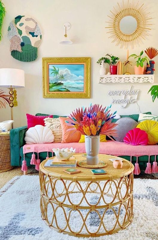 a boho maximalist living room with a green sofa, colorful pillows, rattan tables, bold artworks and a sunburst mirror and potted plants