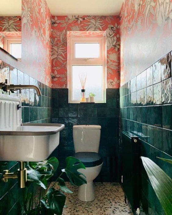a bold maximalist bathroom with red floral wallpaper, dark green tiles, statement plants brass touches