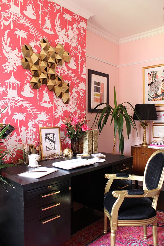 a bold maximalist home office with a bright pink wallpaper wall, a black desk, a refined black and gold chair, cool artworks on the walls