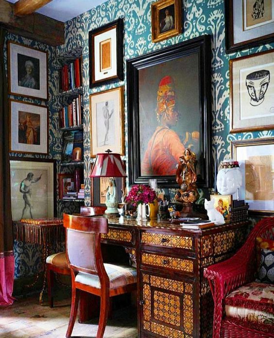 a bold maximalist home office with teal printed wallpaper, gallery wlal on several walls, an inlaid desk, bold printed chairs and a colorful lamp